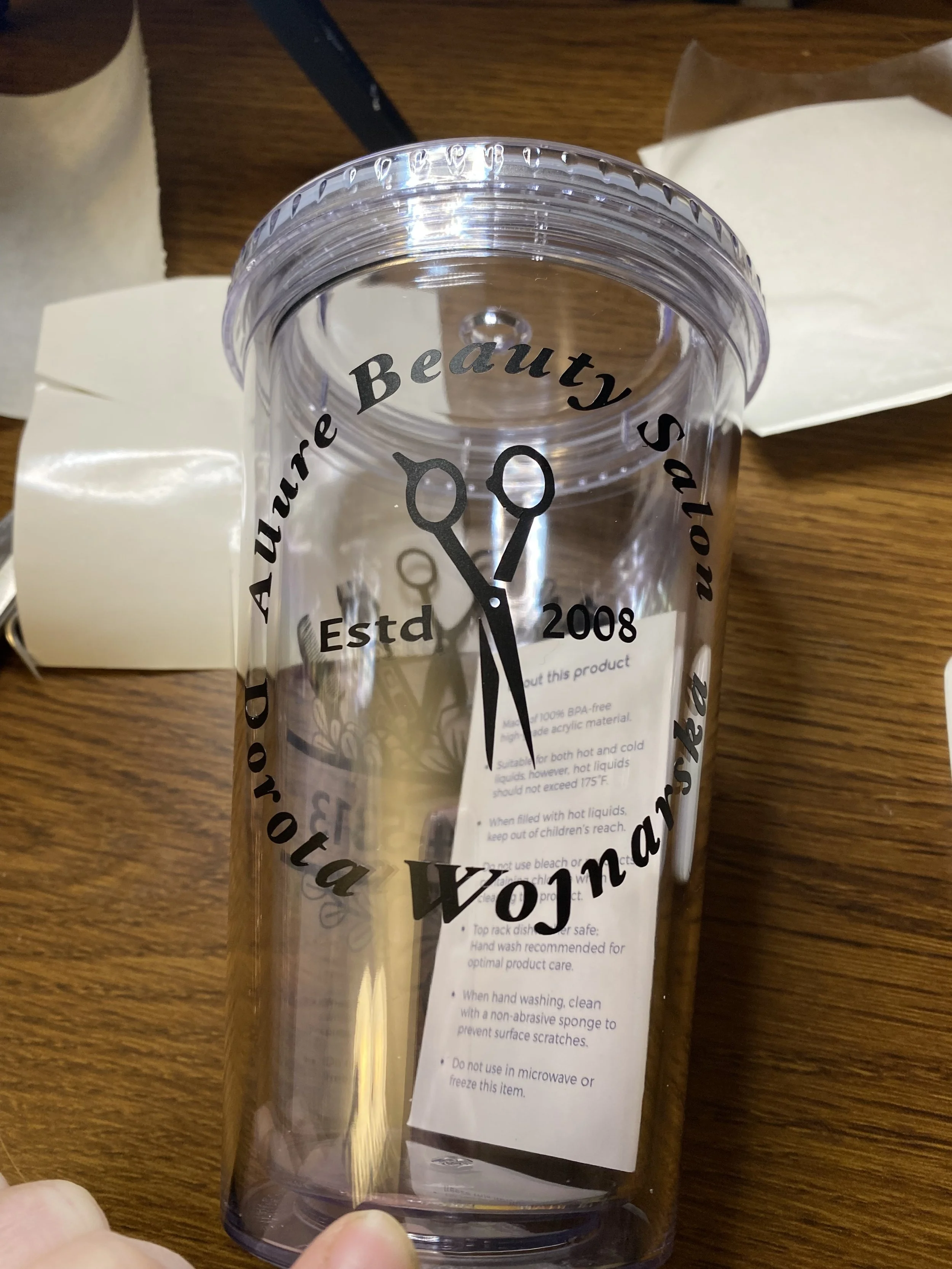 Clear plastic tumbler with black printed text and clock design wrapped around it. The text includes phrases like 'You are aure', 'Founded', 'Estd 2008', and 'Beauty salon'. The tumbler is on a wooden table with papers and a pen visible in the background.