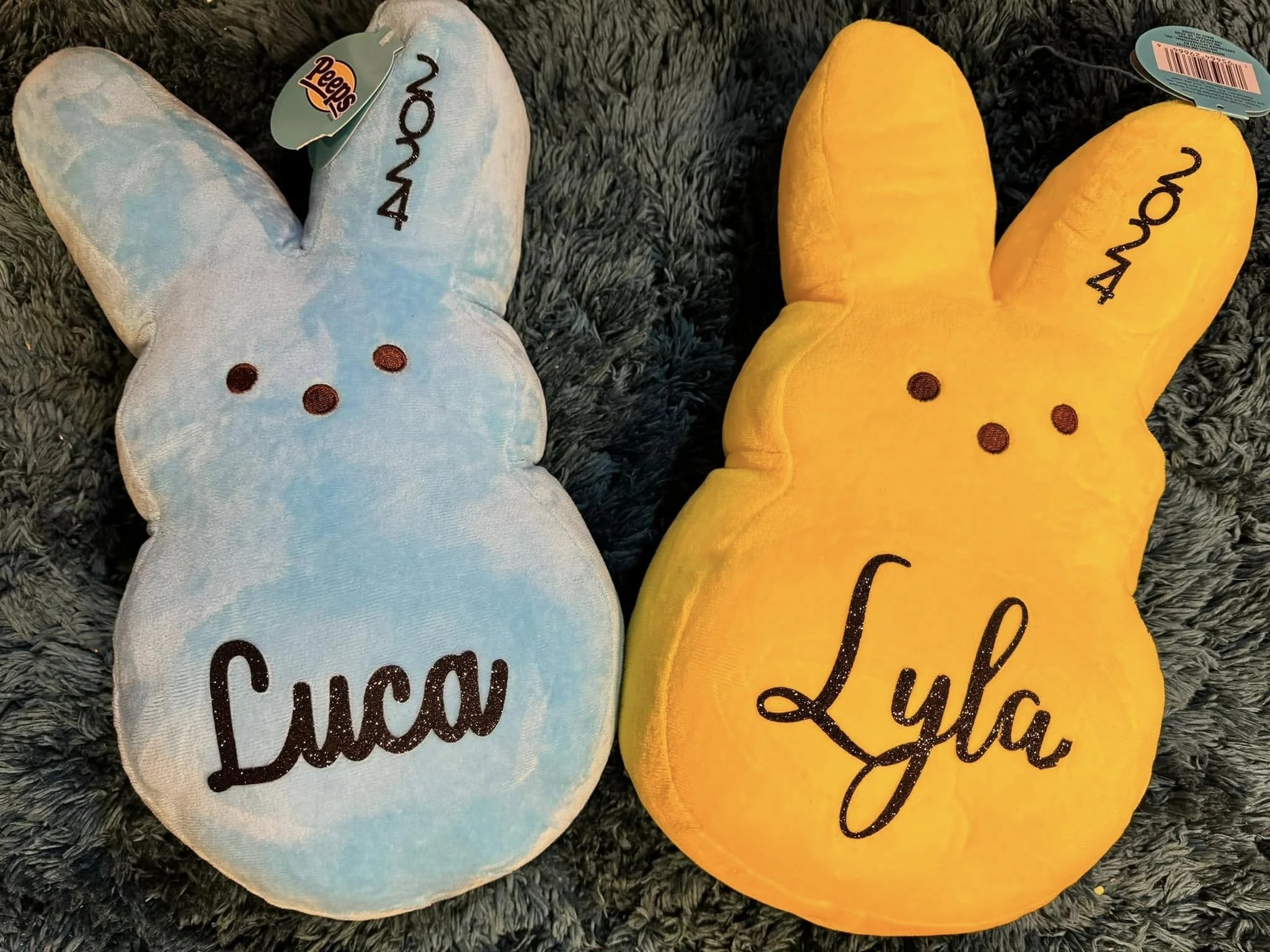 Two plush bunny slippers, one blue with the word 'sweet' embroidered in black and the other yellow with the word 'love' embroidered in black, placed on a gray textured surface.
