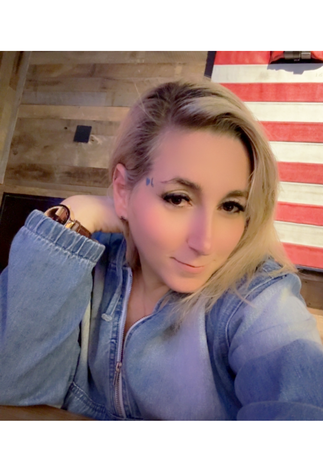 A woman with blonde hair in a denim jacket is sitting indoors, resting her head on her hand. Behind her, there is a wall with wood paneling and a red and white striped decorative piece. She is smiling softly and looking at the camera.