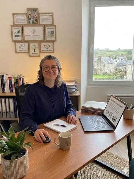 Femme souriante assise à un bureau avec ordinateur portable, mug, plantes et étagères de livres dans un bureau avec fenêtre.