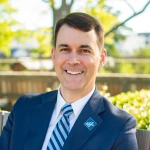 A smiling man in a suit with a blue tie and a lapel pin, outdoors with a wooden fence and trees in the background.