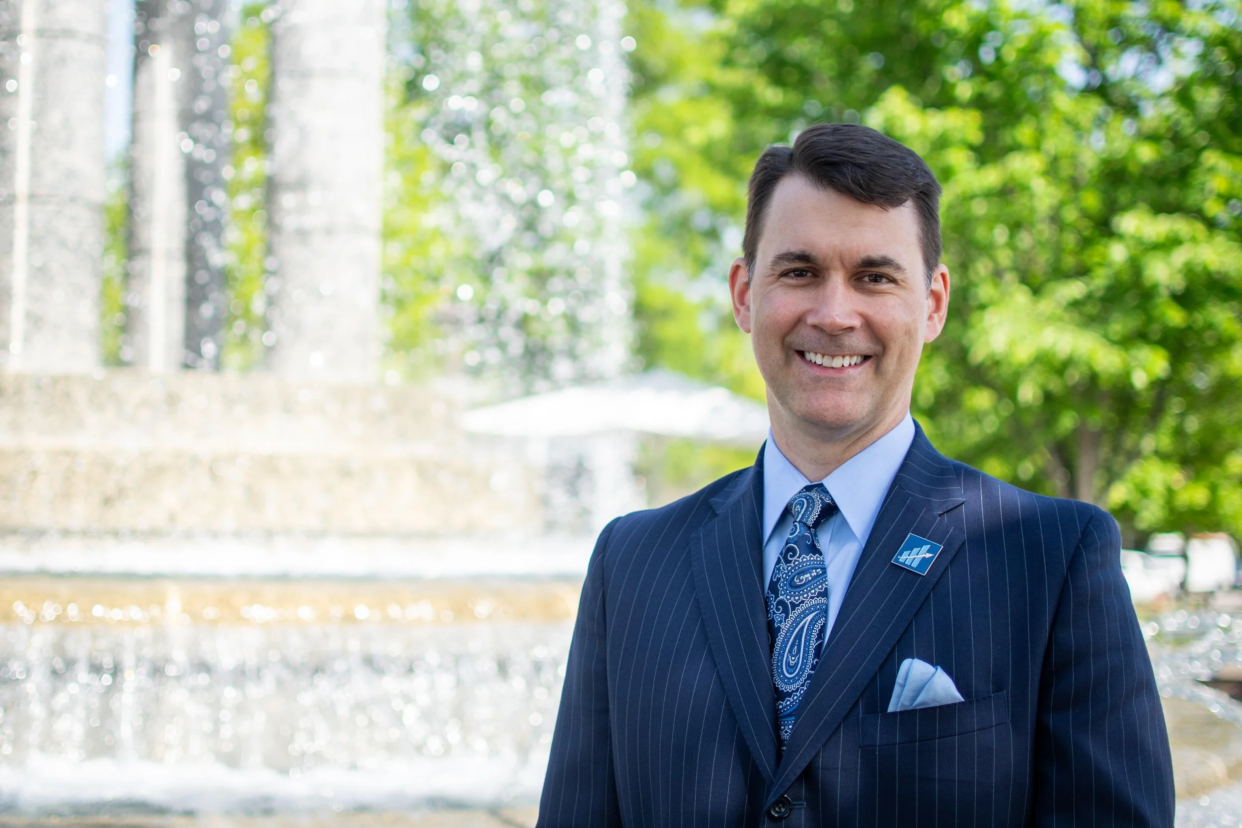 A smiling man wearing a pinstripe suit, light blue shirt, and patterned blue tie standing outdoors in front of a fountain and green trees.