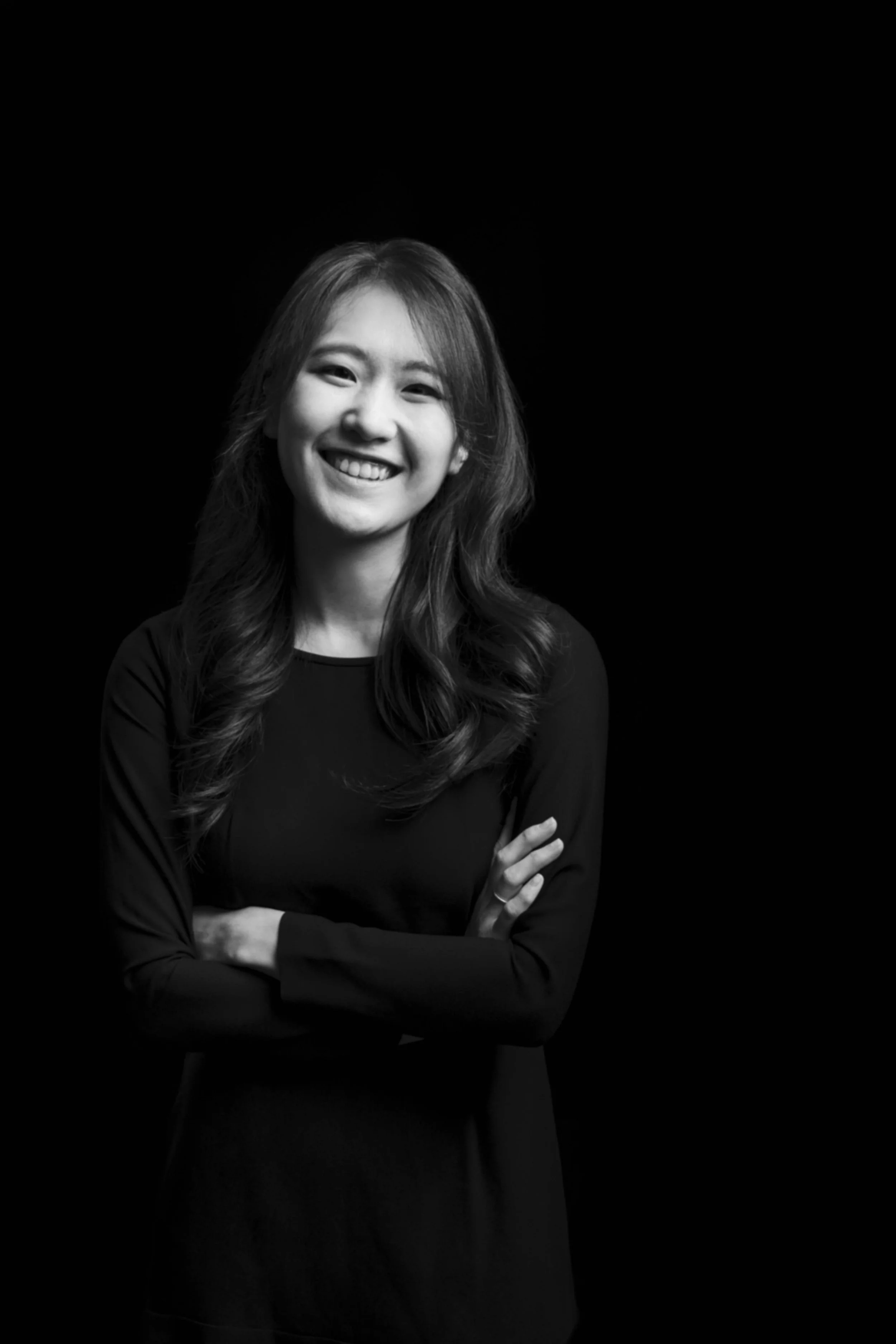 Black and white portrait of a smiling woman with long wavy hair, crossing her arms, against a dark background.