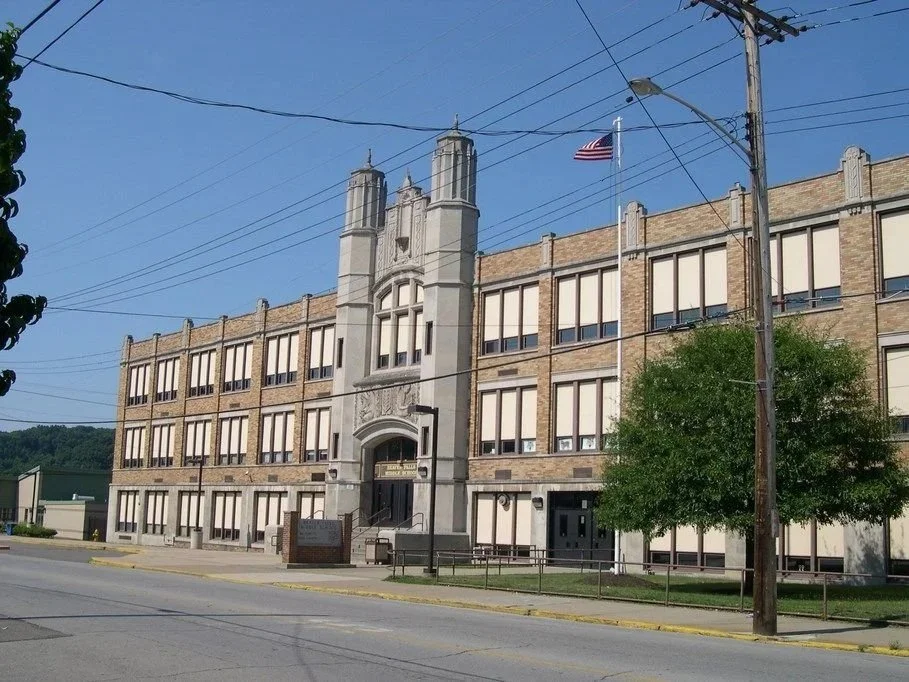 A school building with a brick exterior, arched entrance, and four tall towers above the entrance, located on a street with power lines, a flagpole with an American flag, and a tree in front.