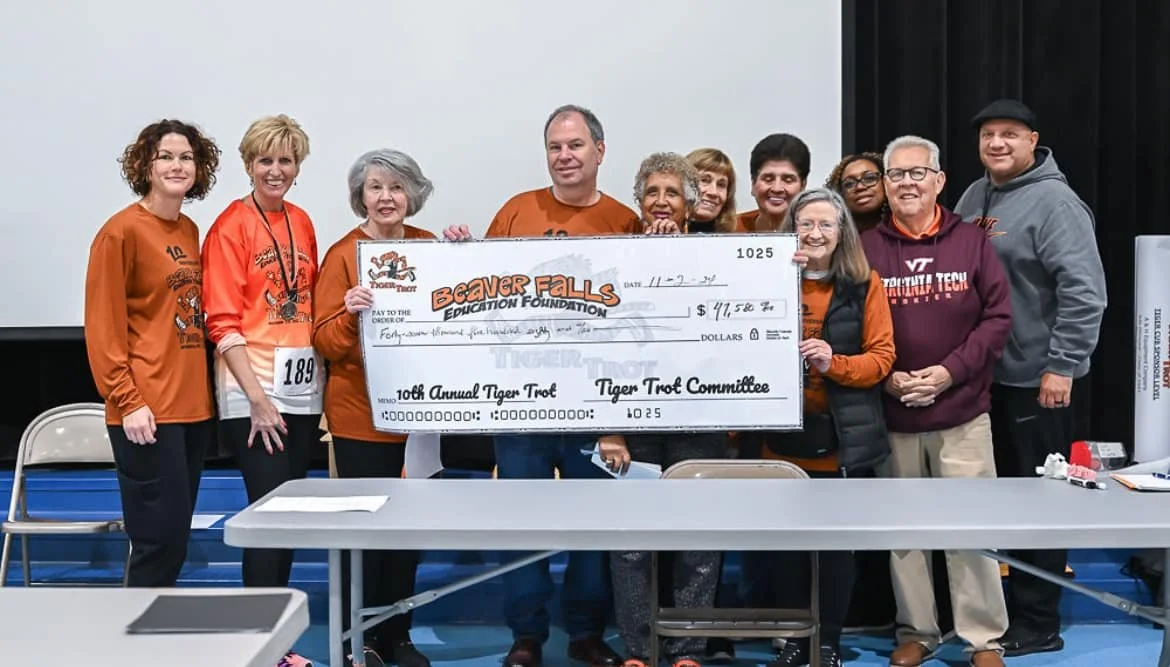 Group of people holding a large check for $41,580 during the 10th Annual Tiger Trot event at Beavers Falls, with some people wearing orange and others in casual clothing, standing in front of a white and black background.