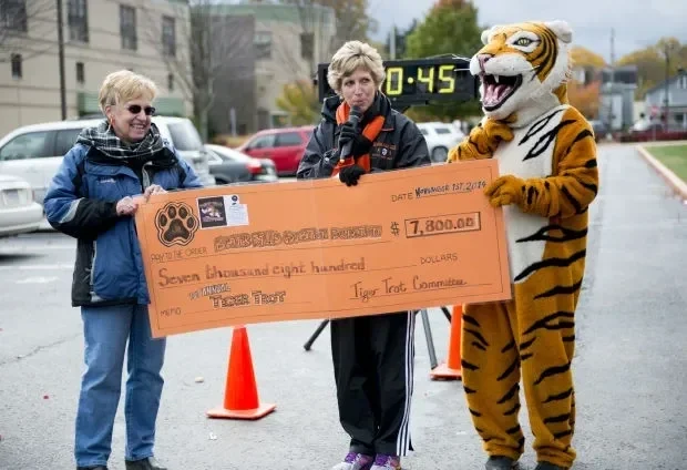 Two women and a person in a tiger costume holding a large check for $7,800 at an outdoor charity event.