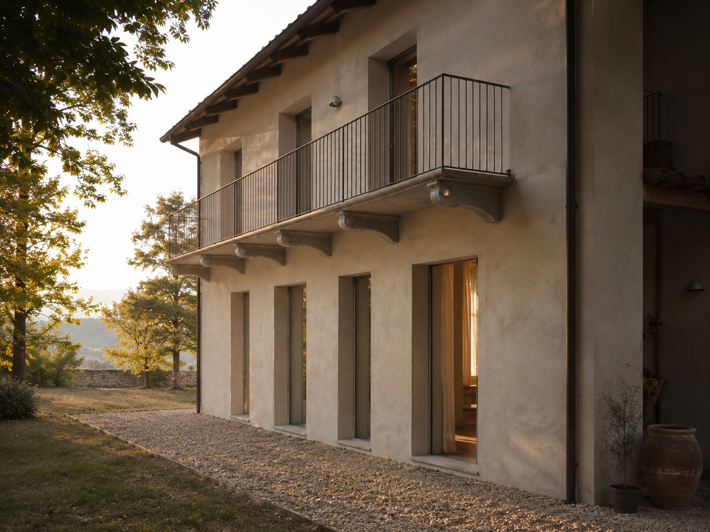 A two-story house with a beige stucco exterior, large vertical windows, and a metal balcony railing on the second floor. The house is surrounded by trees and sits in a natural landscape at sunset.
