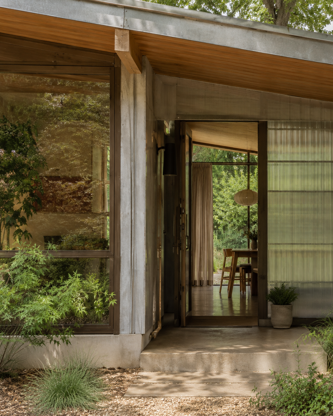 Open doorway showing a cozy interior with a table, chairs, a floor lamp, and greenery outside, viewed through textured glass panels.