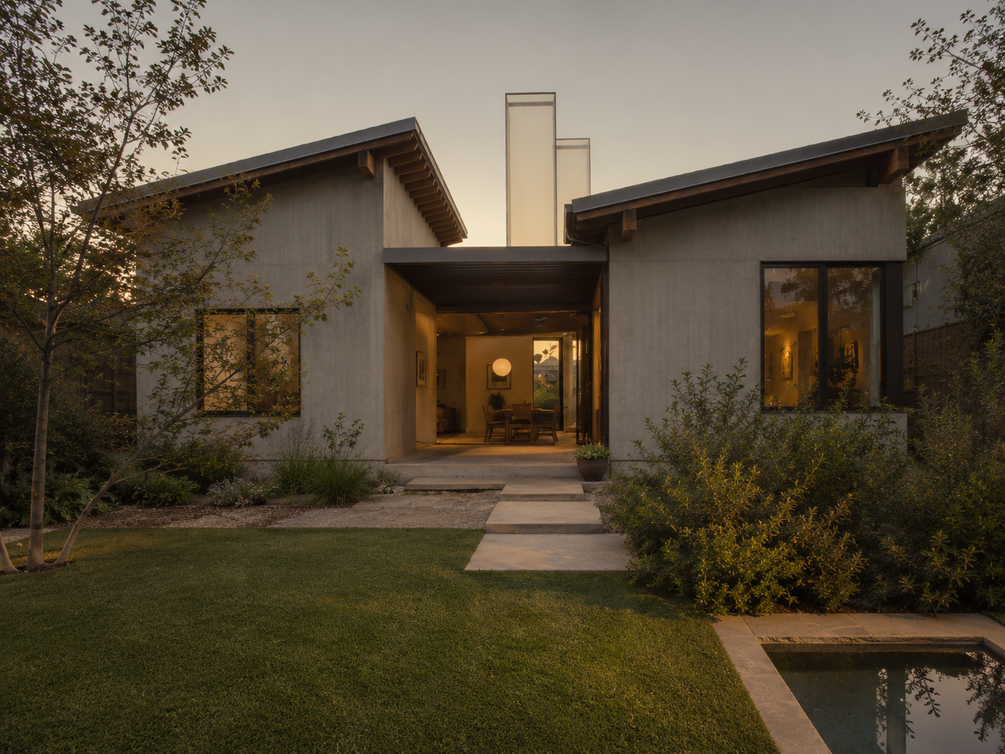 A modern house with large windows, a small front garden, and a concrete pathway leading to the entrance. The house has a minimalist design and is illuminated from inside, with trees and greenery around.