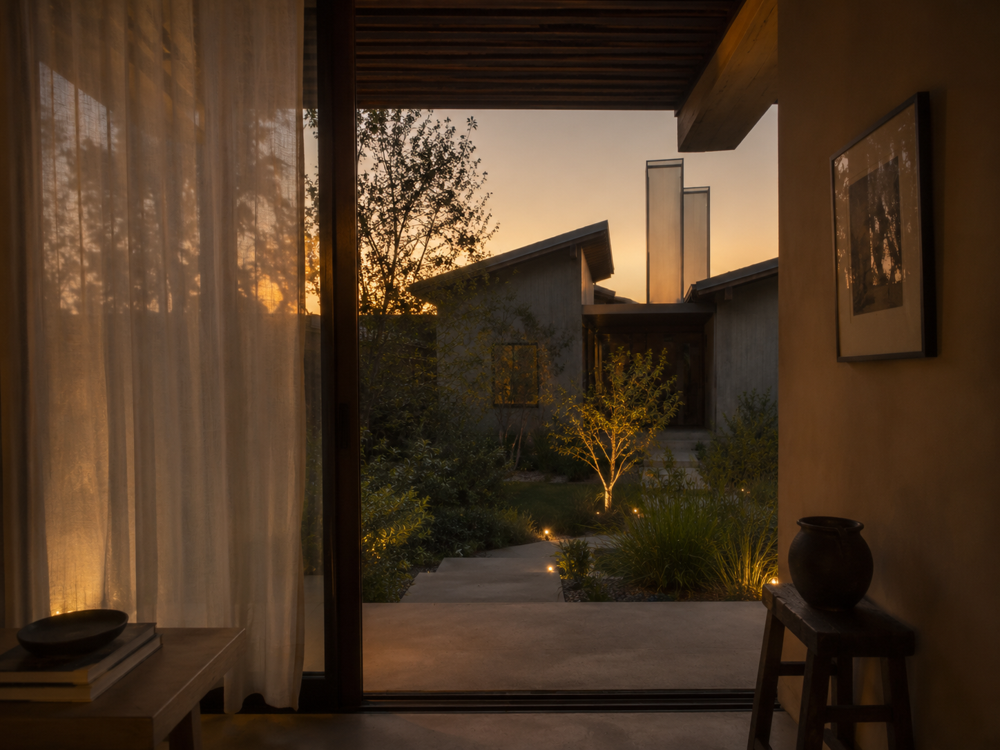 View of a modern house's backyard at sunset, seen from inside through a sliding glass door with sheer curtains. The backyard has small trees, shrubs, and a stone pathway with outdoor lighting.