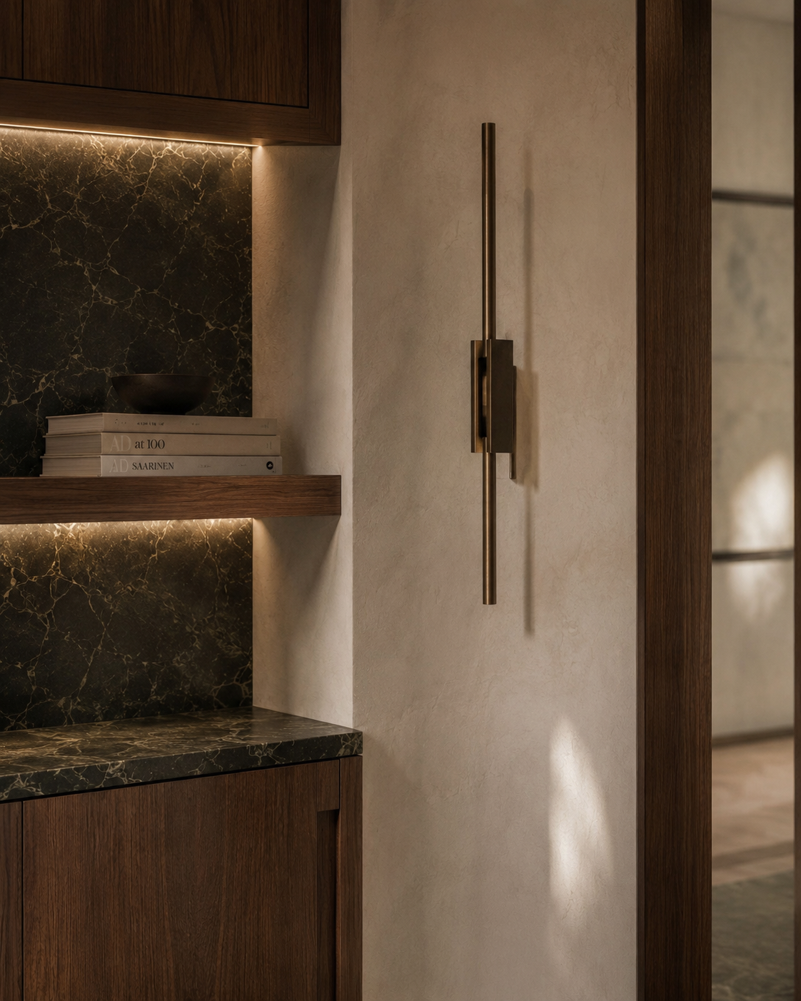 Interior view of a modern living space showing a wall with a vertical metallic wall lamp, a shelf with books and a bowl, and a dark marble countertop with wooden cabinetry.
