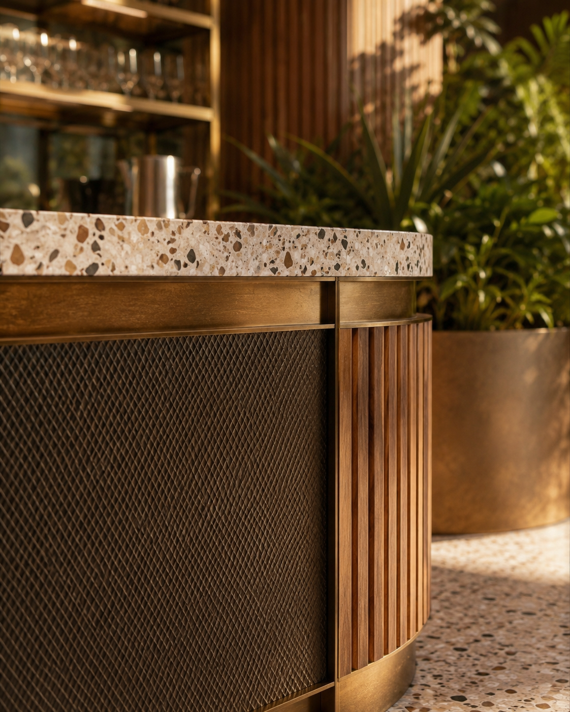 Close-up of a bar counter with a marble top and textured wood and metal facade, background with green plants and wooden shelves with glasses.
