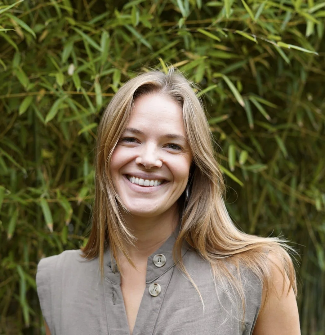 A woman with long light brown hair smiling outdoors in front of green bamboo plants.