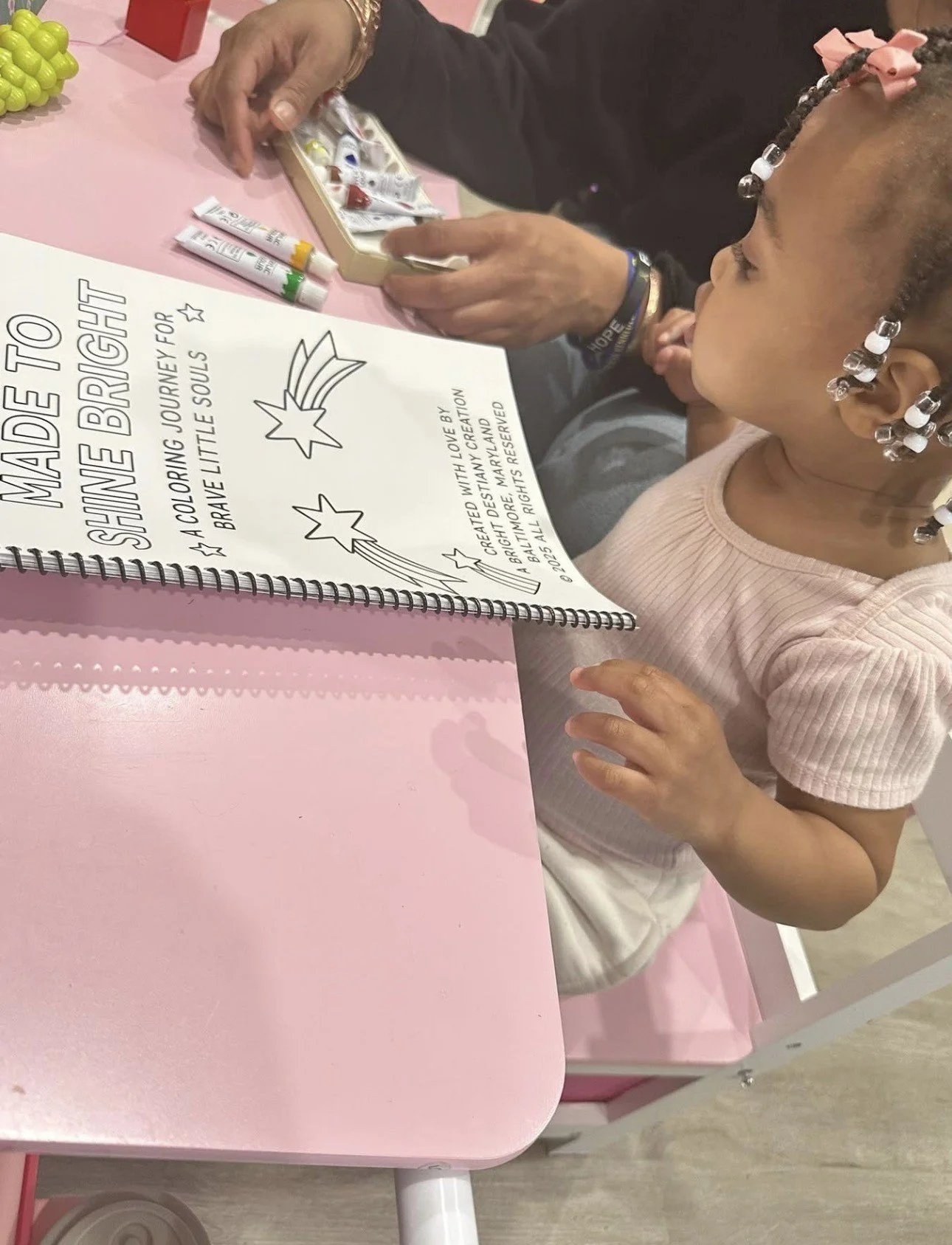 A young girl with bead earrings and a pink hair bow is sitting at a pink table, looking at a coloring book. An adult is sitting next to her, holding a box of paints. The coloring book has the words 'Made to Shine Bright' on the cover.