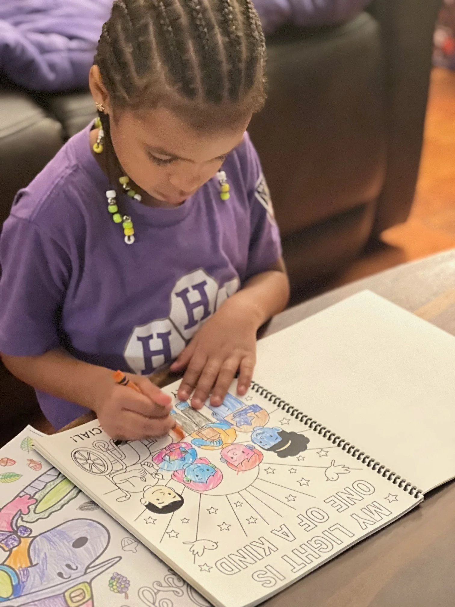 A young girl with braided hair and beads is coloring a coloring page with illustrations of girls, animals, and hearts. The coloring page has a large circle with the words 'MY LIGHT IS' and 'ONE OF A KIND' visible. The girl is sitting at a table in a room with a couch and pillows in the background.