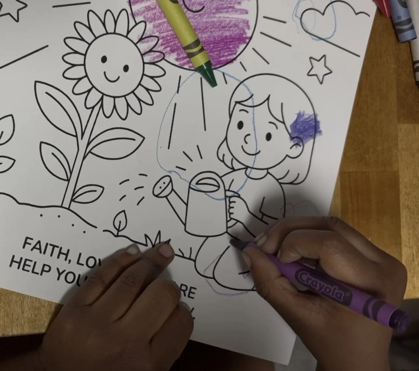 A person uses a purple Crayola marker to color a children's coloring page featuring a girl watering a sunflower with a smiling sun and clouds in the background.