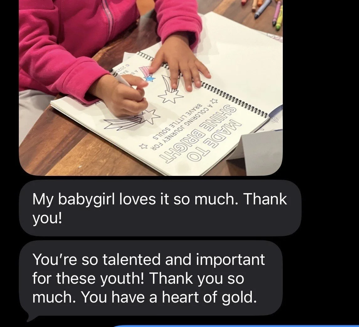 A child in a pink jacket coloring a patriotic page with stars and a rocket on a wooden table, with crayons scattered around, next to an open blank notebook.