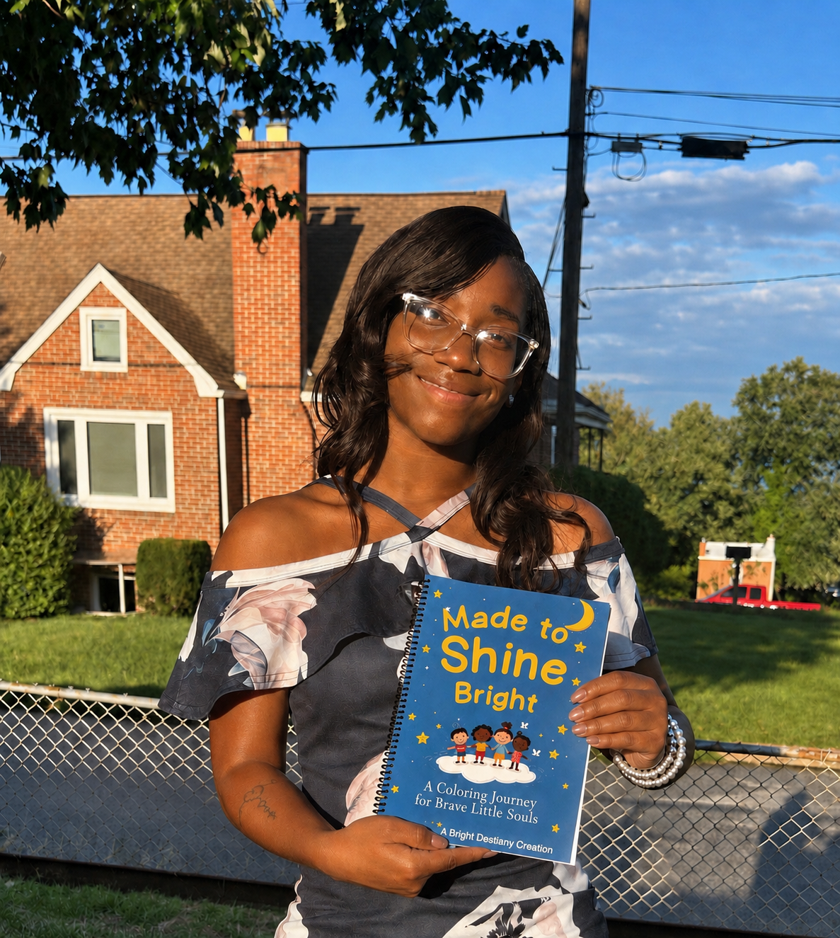 A young woman with glasses smiling while holding a colorful coloring book titled 'Made to Shine Bright' outdoors in front of a house with brick walls, trees, and a blue sky.