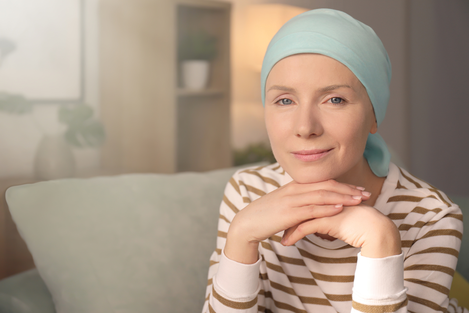 A woman with light skin and blue eyes wearing a light blue headscarf and a beige and white striped shirt, sitting on a couch in a cozy, well-lit room.