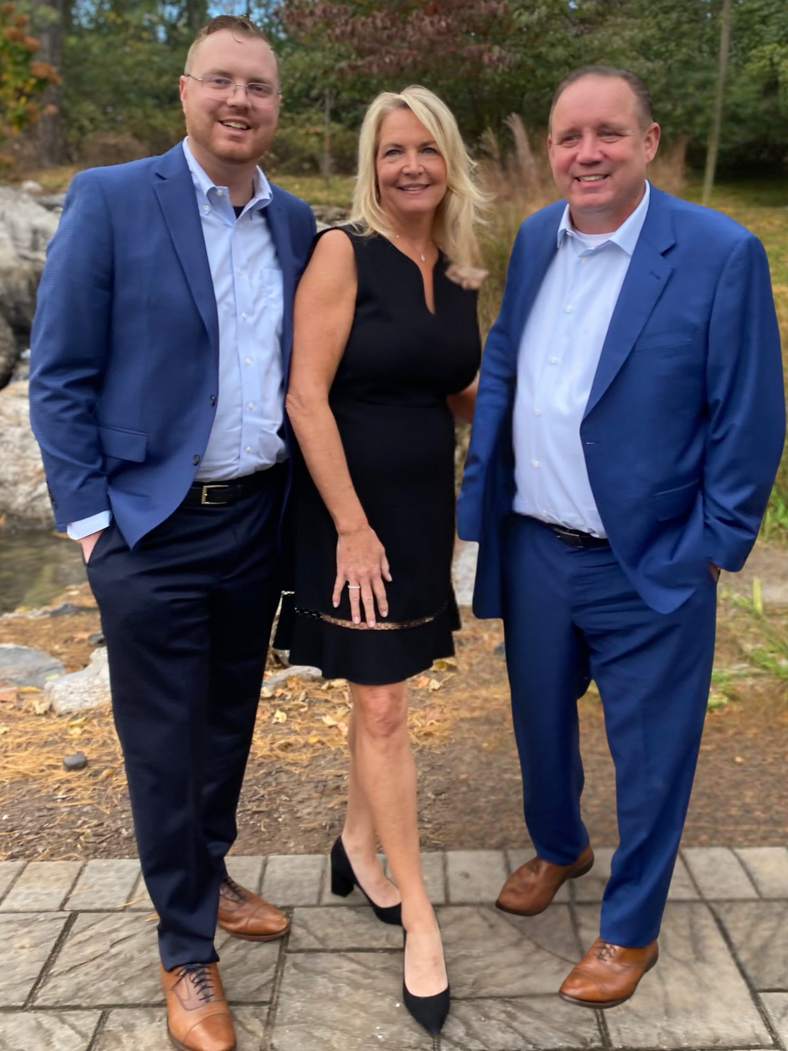 Three people dressed in formal attire standing outdoors on a paved path, with trees and fall foliage in the background.