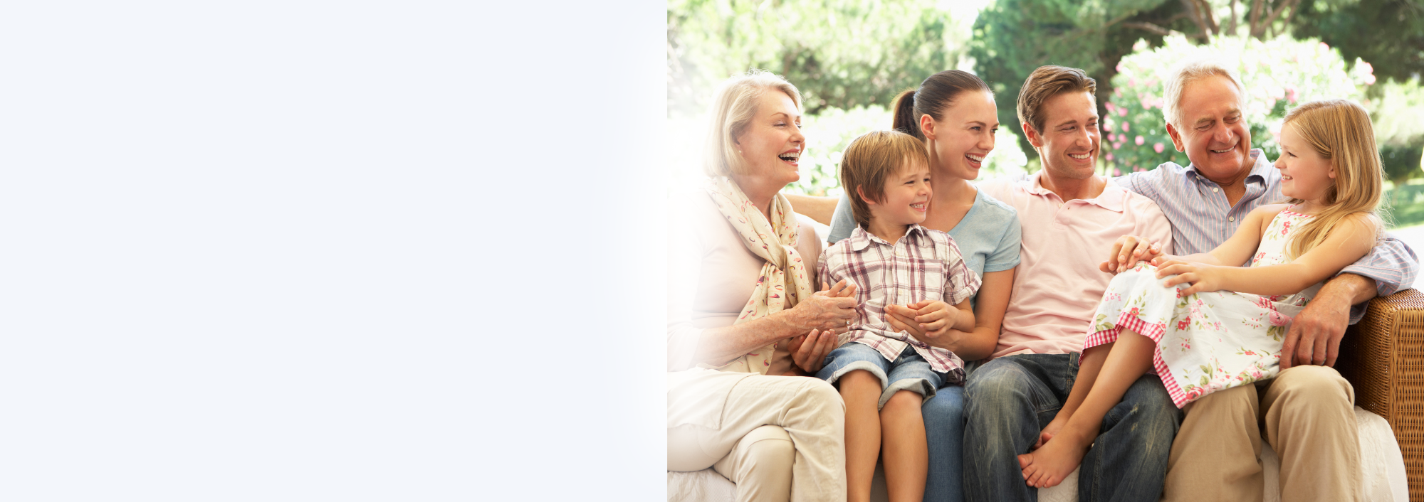 Family of six, including two children, sitting on a outdoor sofa, smiling and laughing together on a sunny day with trees and flowers in the background.