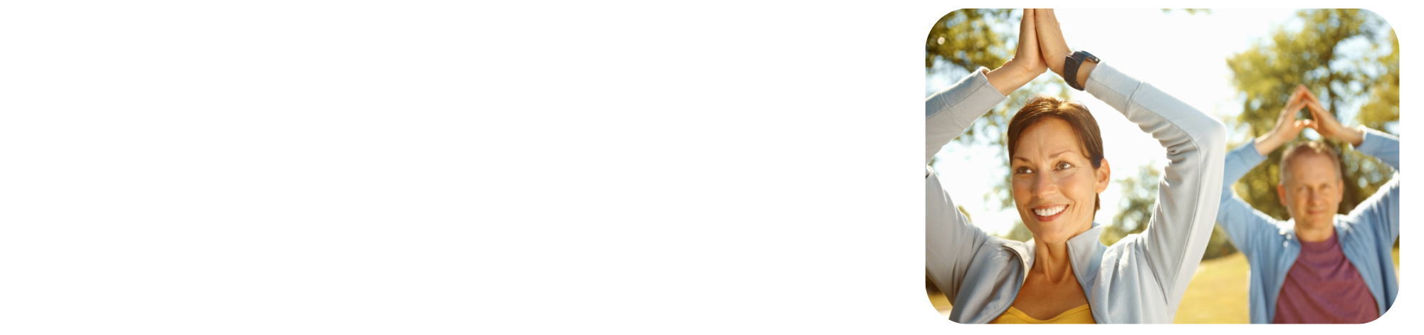 Woman and man practicing yoga outdoors with hands together above their heads, smiling on a sunny day with trees in the background.