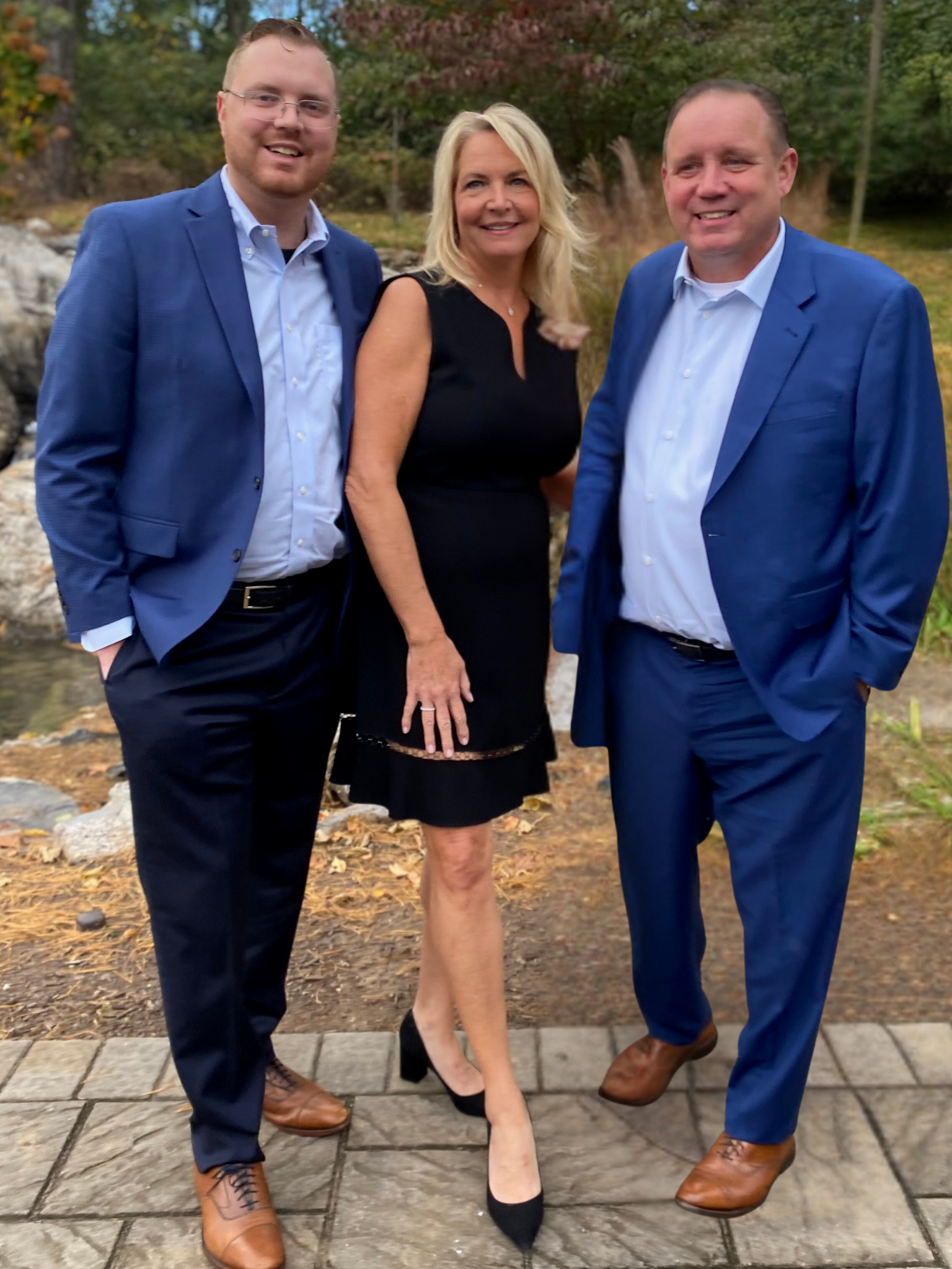 Three people standing outdoors on a paved path, dressed in formal attire, with trees and autumn foliage in the background.