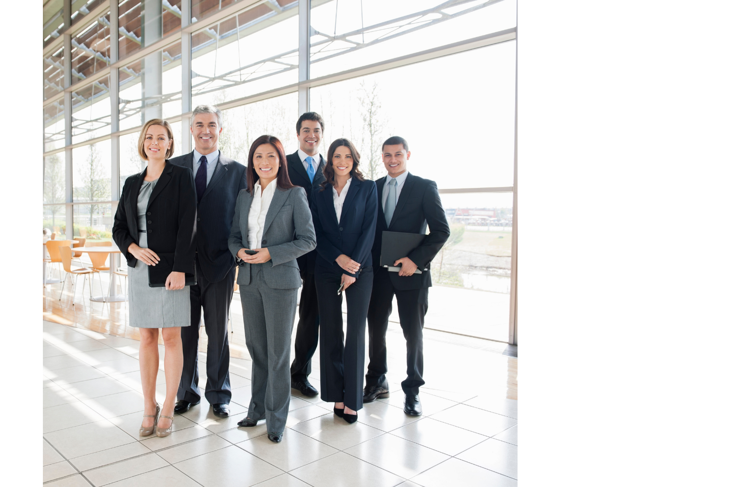 Group of six diverse business professionals standing in a modern office lobby with large windows and natural light.