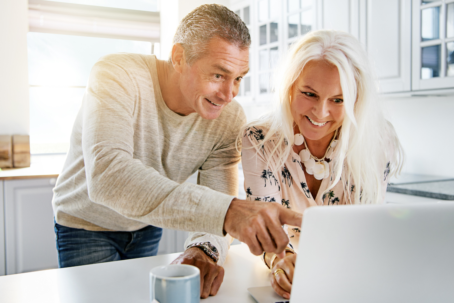 A senior man and woman smiling and looking at a laptop on a kitchen countertop, with the man pointing at the screen and the woman using a mouse, in a bright kitchen with white cabinets and large windows.