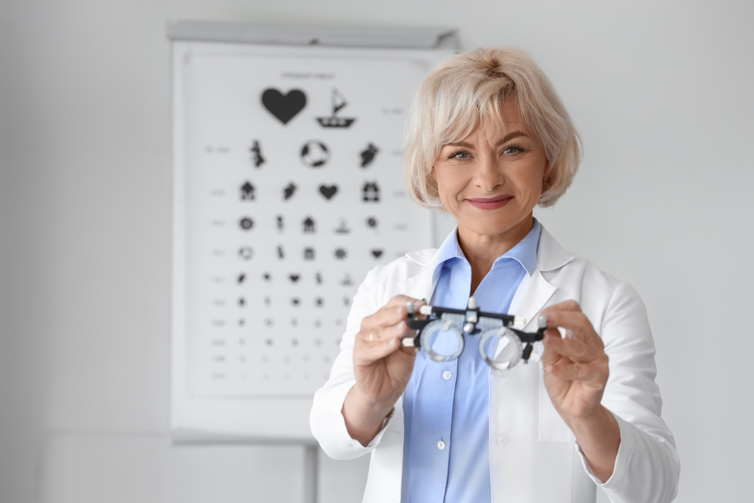 Optometrist smiling and holding a trial frame in an eye clinic with an eye chart in the background.