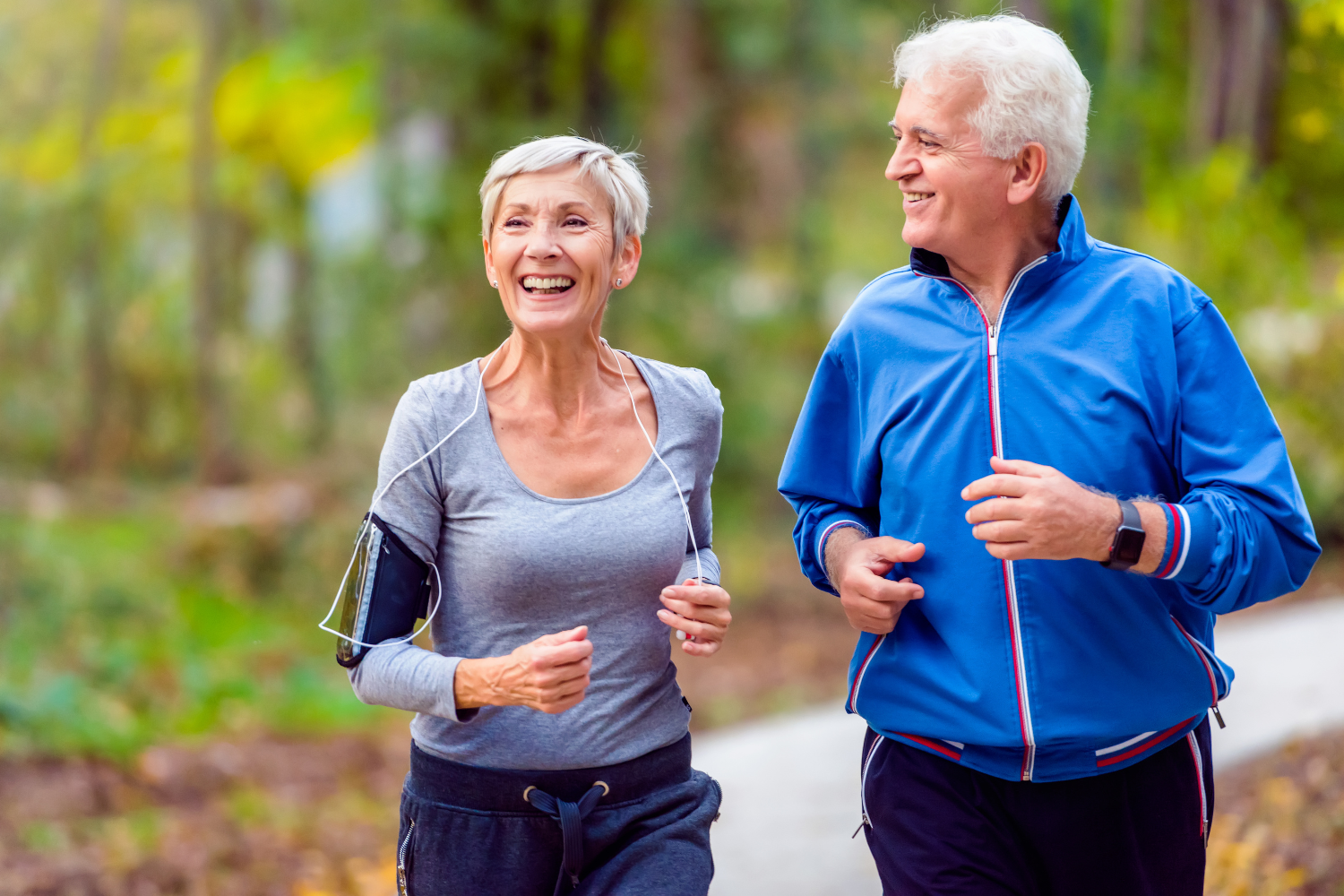 An elderly couple jogging outdoors on a forest trail, smiling and enjoying their exercise.