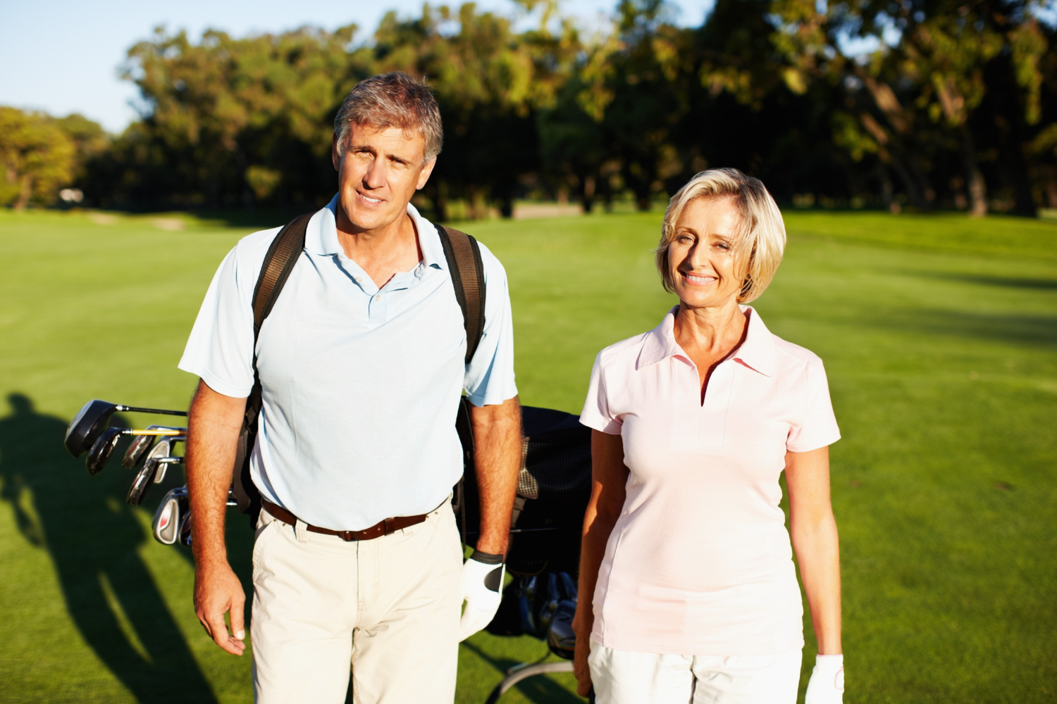 A mature man and woman standing on a golf course, smiling, with golf clubs and a golf bag. The man is wearing a light blue polo shirt and khaki pants, and the woman is wearing a pink polo shirt and white pants. There are trees and a bright sky in the background.