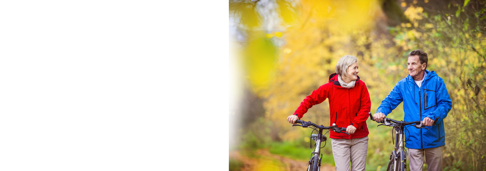 An elderly man and woman walking their bikes on a forest trail, smiling and looking at each other, surrounded by yellow autumn leaves.