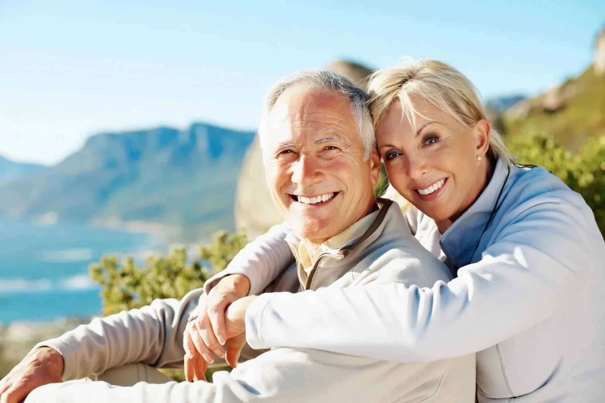 An elderly couple smiling outdoors near a body of water with mountains in the background.
