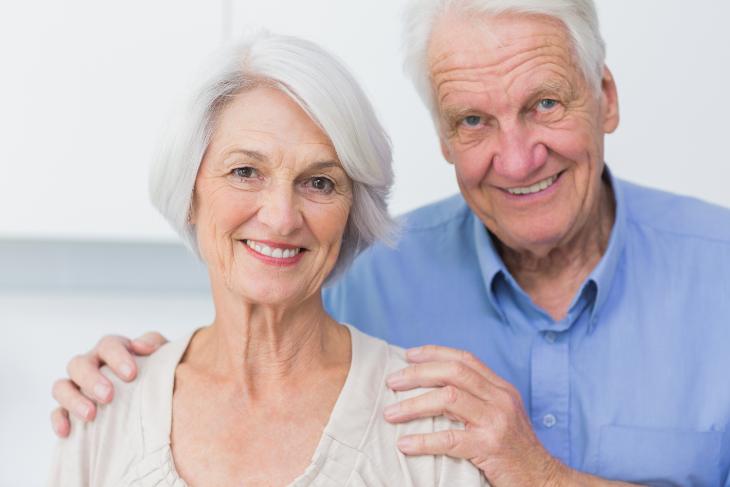 An elderly couple smiling and standing close together, with the man's hand resting on the woman's shoulder.