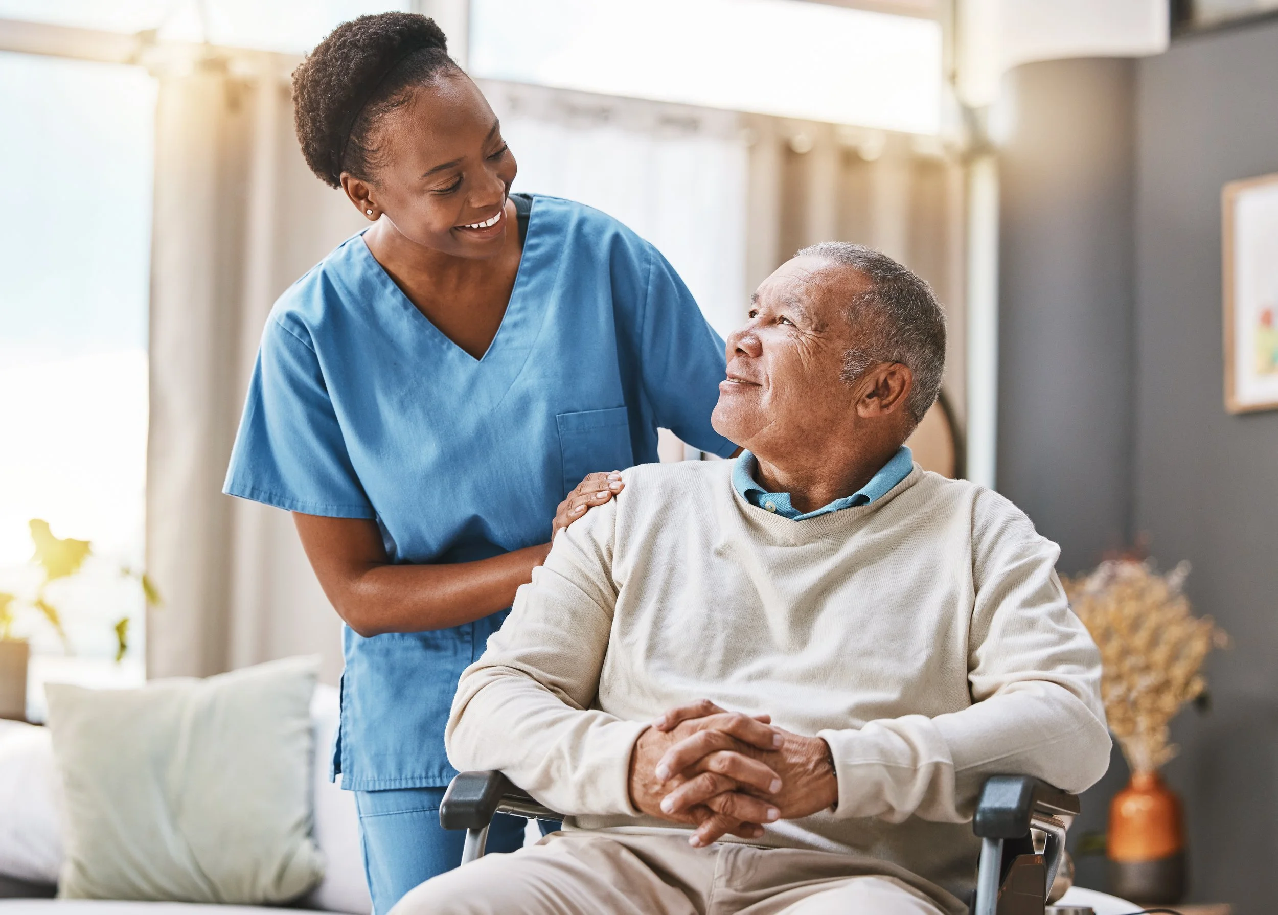 A young female healthcare worker with dark curly hair, wearing blue scrubs, smiling and talking to an elderly man in a wheelchair, in a bright medical facility.