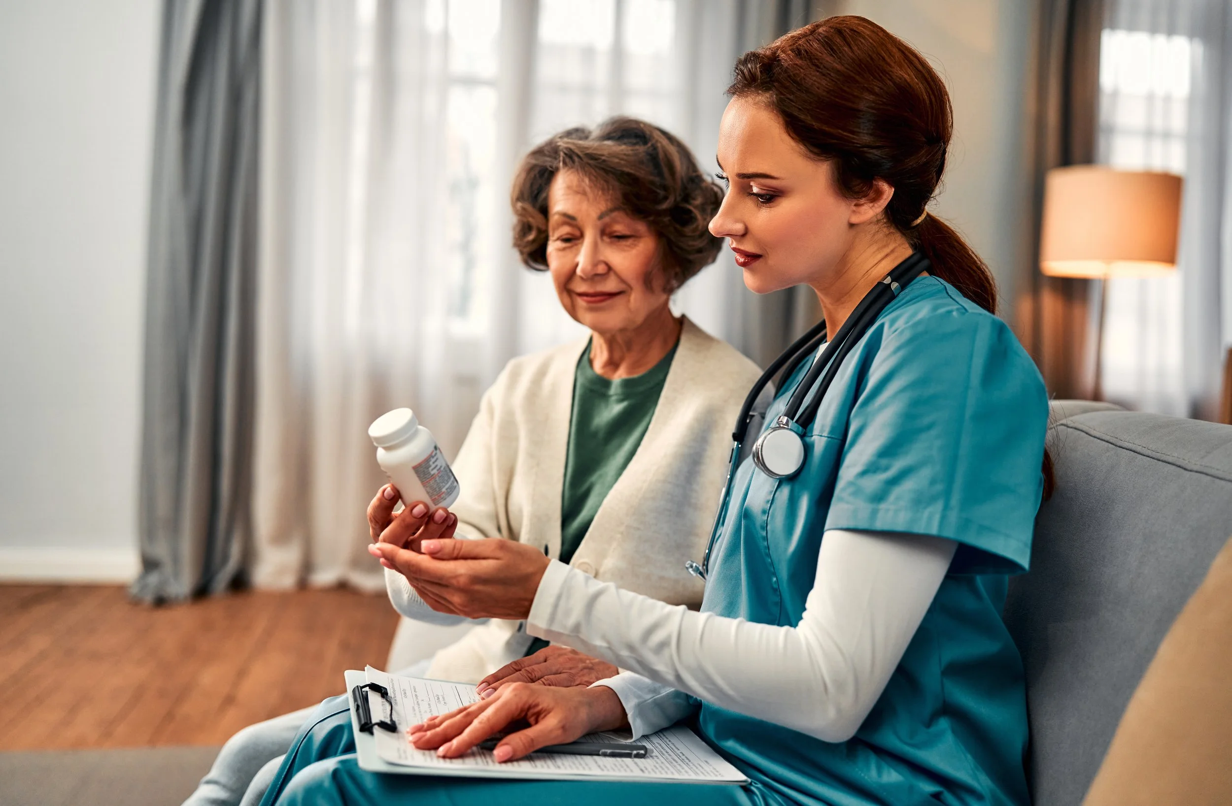 A healthcare professional, wearing blue scrubs and a stethoscope, sitting beside an elderly woman in a bright room with curtains and a lamp, holding a prescription bottle and a clipboard.