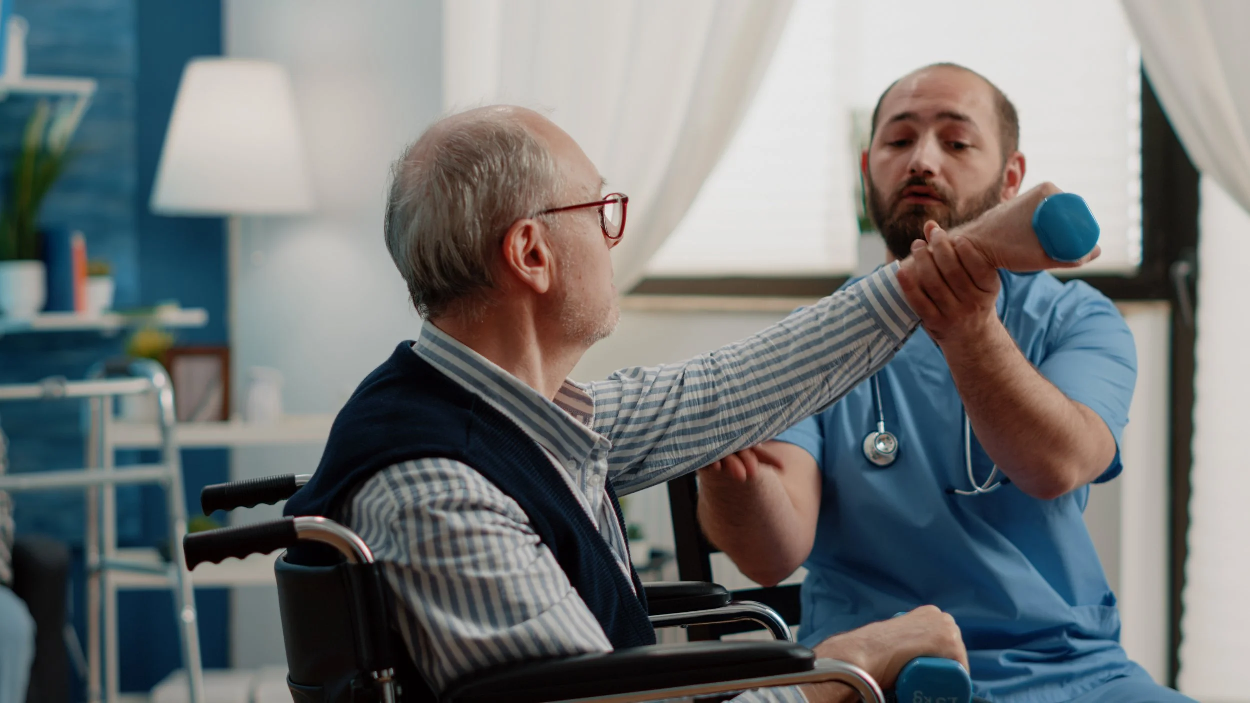 A healthcare professional in blue scrubs assisting an elderly man in a wheelchair by checking his arm with a light blue dumbbell in a medical setting.