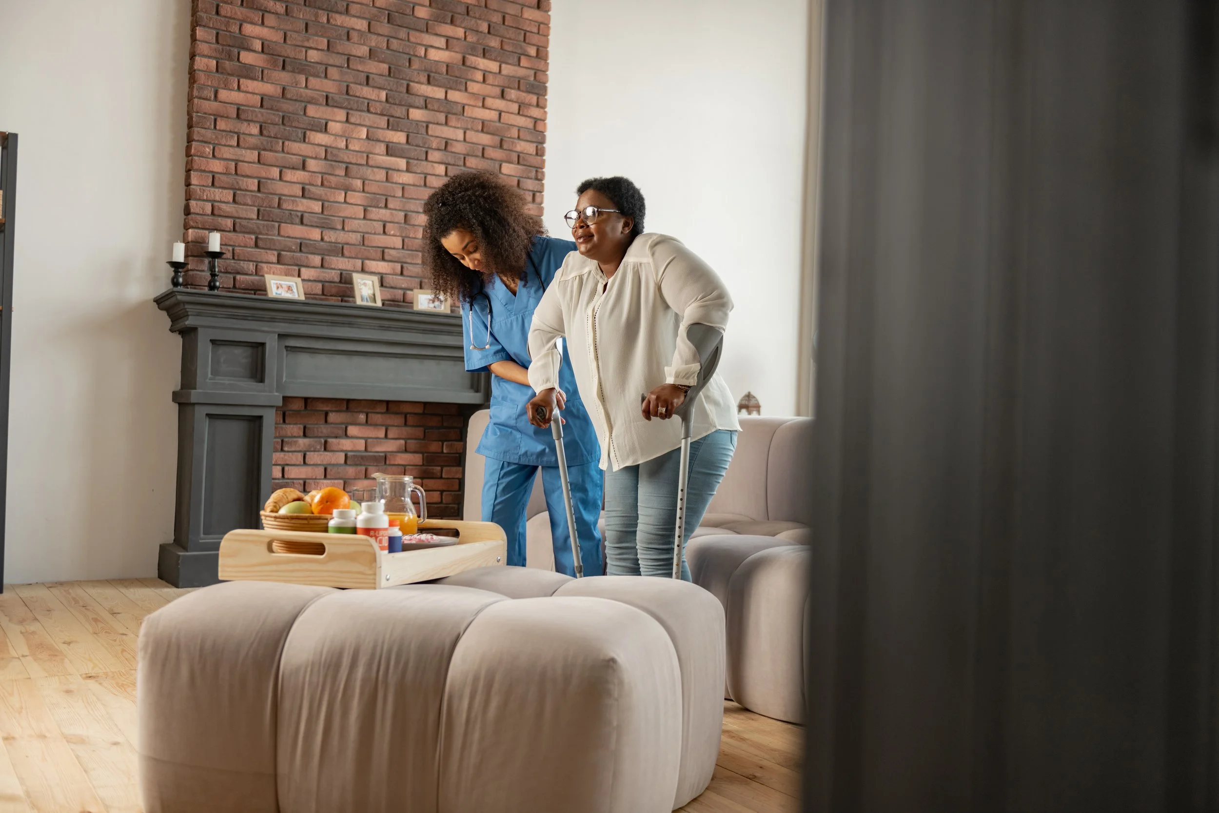 A caregiver helps an elderly woman walk with crutches inside a living room, with a fireplace, framed photos, and a table with fruit and drinks visible in the background.