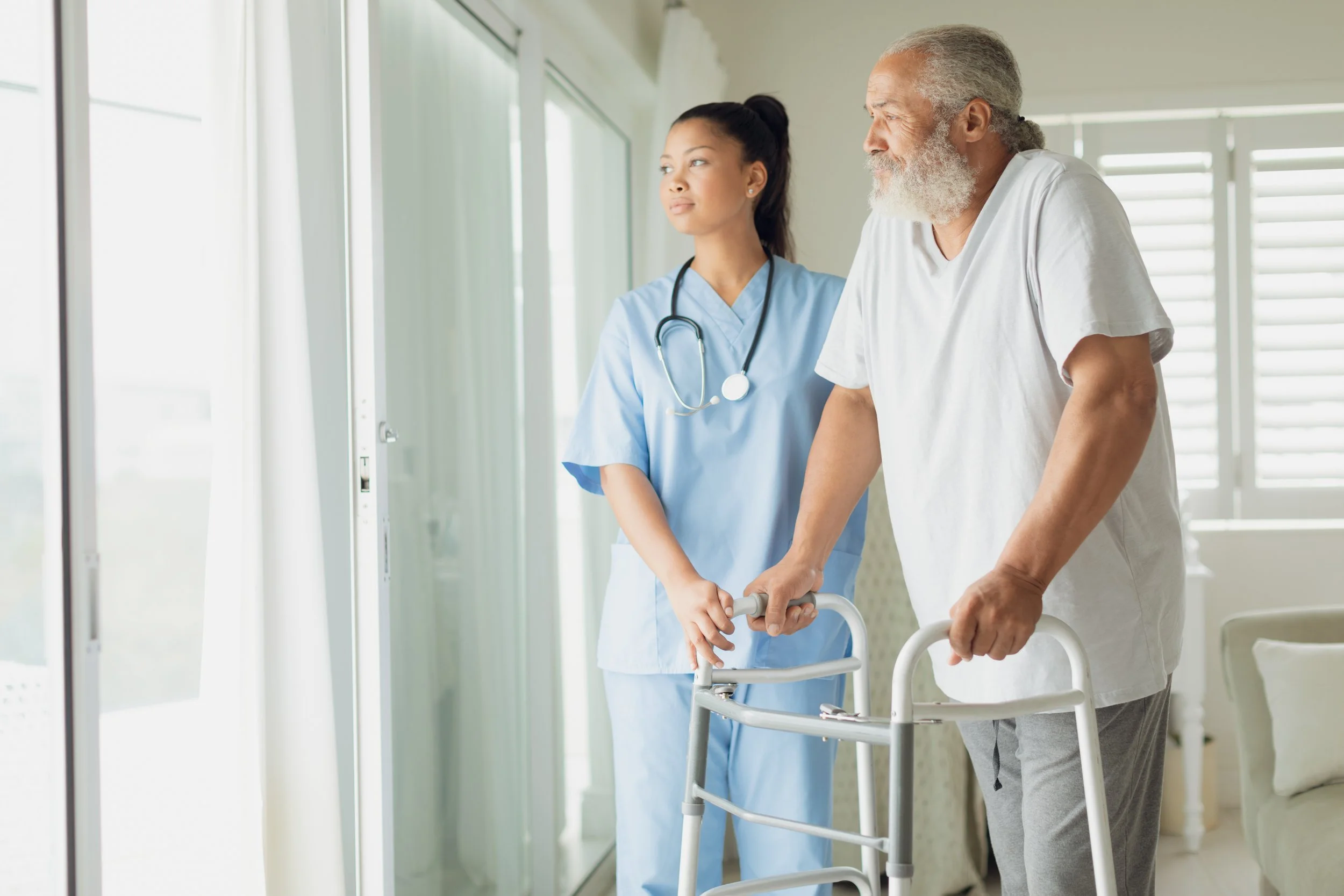 A young female nurse assisting an elderly male patient with a walking aid in a brightly lit healthcare facility.