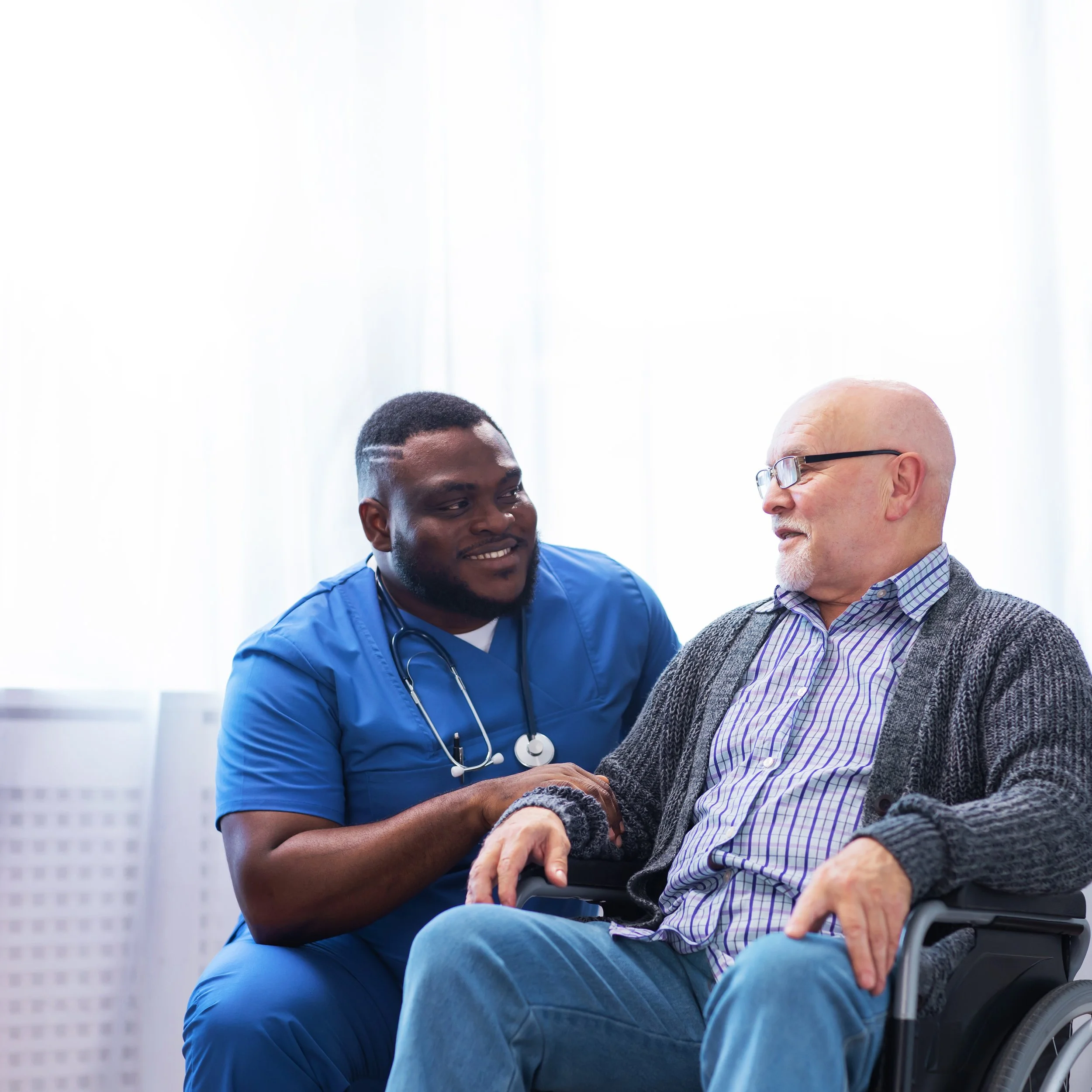 Two men, one in a wheelchair, are engaging in conversation outdoors with a cityscape in the background.
