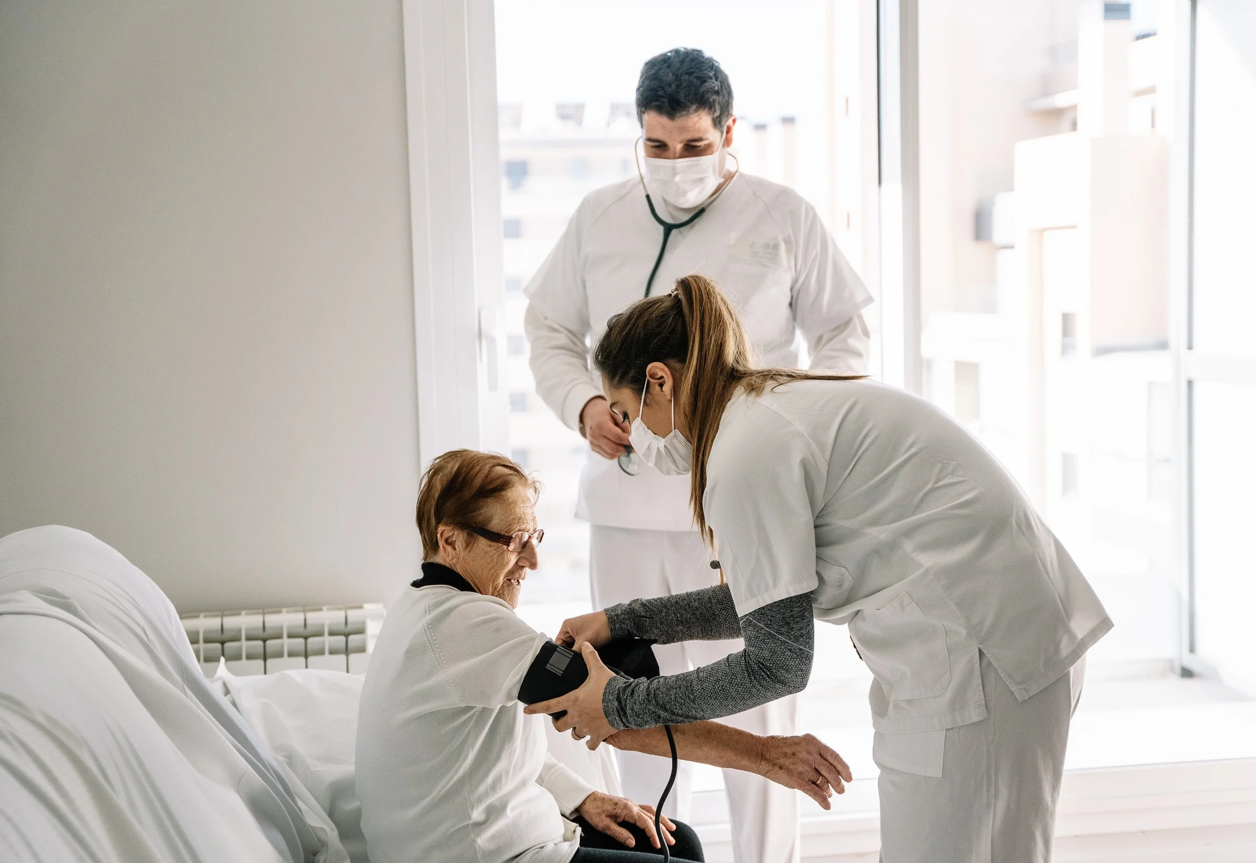 Healthcare professionals in masks attending to an elderly patient in a hospital room with large windows.