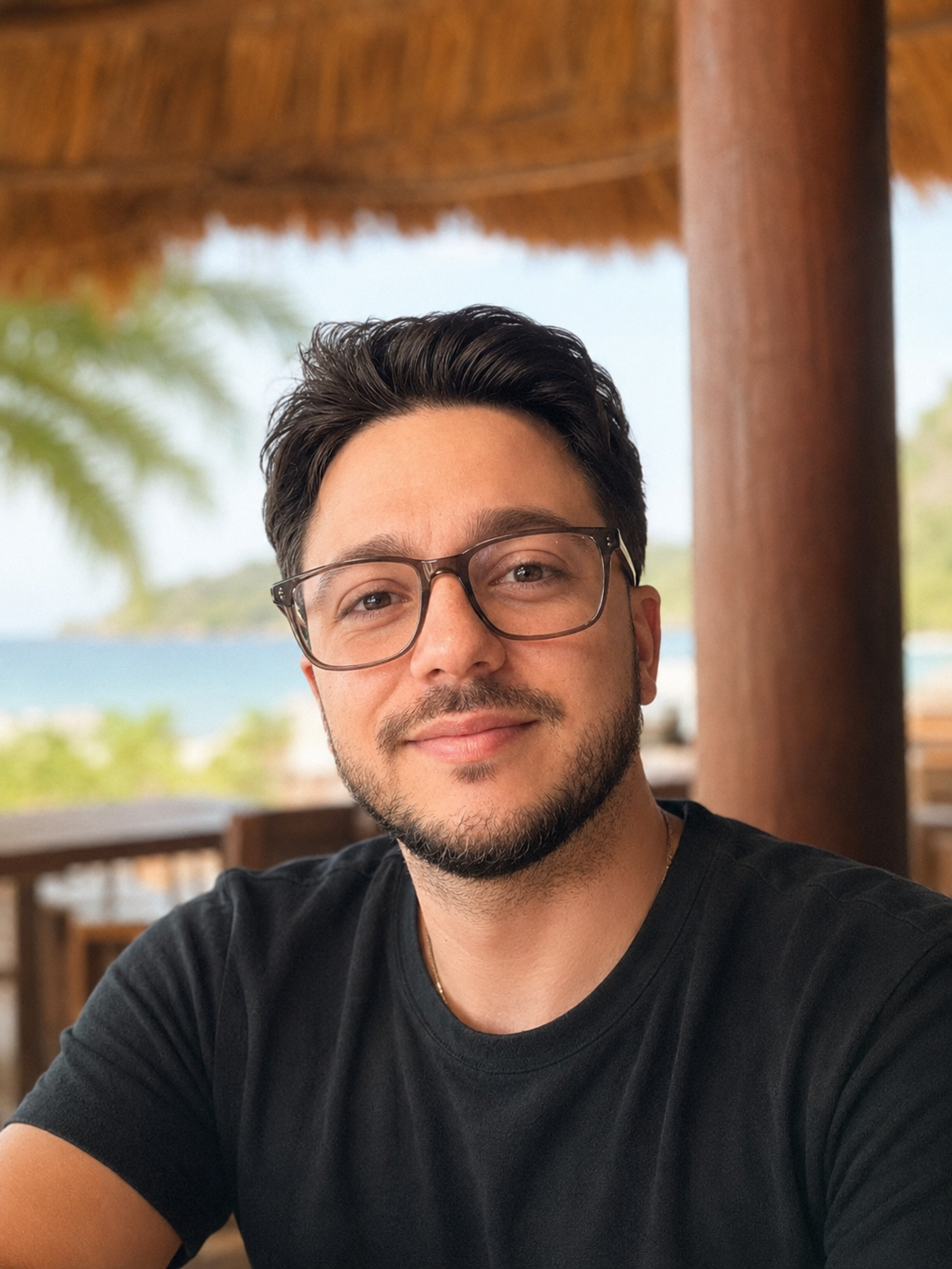 Un jeune homme portant des lunettes souriant, assis dans un lieu en plein air avec un toit en toit de chaume, des arbres et la mer en arrière-plan.