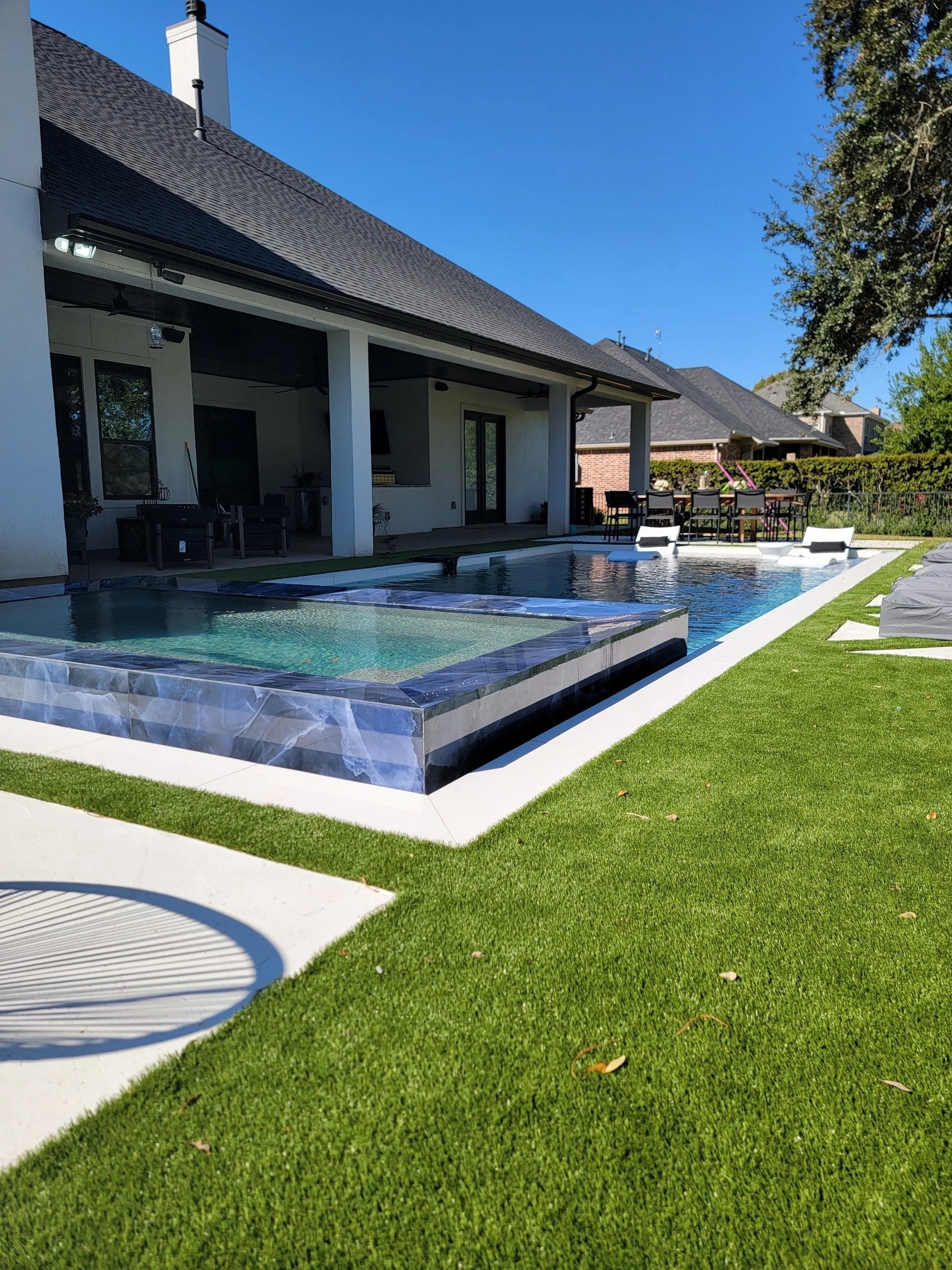Backyard view with a swimming pool, outdoor furniture, and a patio area, with a house and trees in the background under a blue sky.