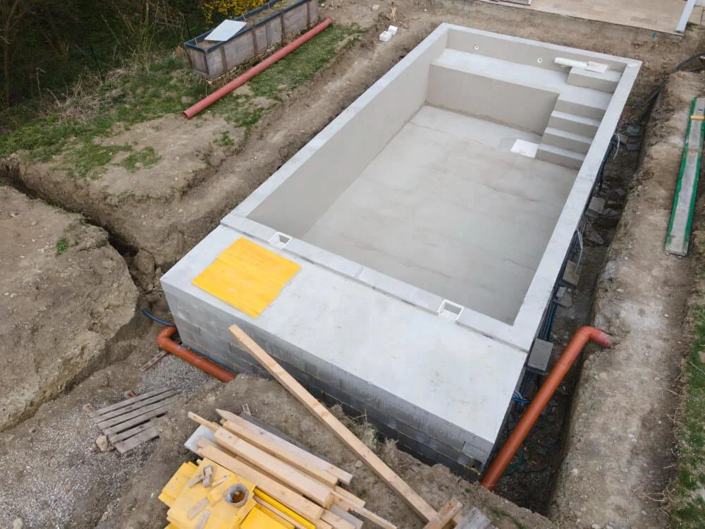 Empty swimming pool under construction in a backyard, with stairs at one end and plumbing installed around it.