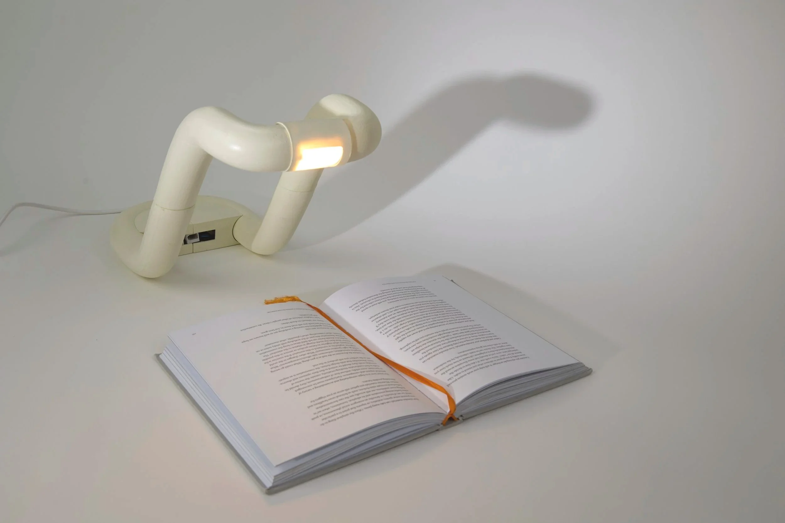 An open book with a bright orange ribbon bookmark lying on a white surface, with a beige curved desk lamp above casting a shadow on the wall.