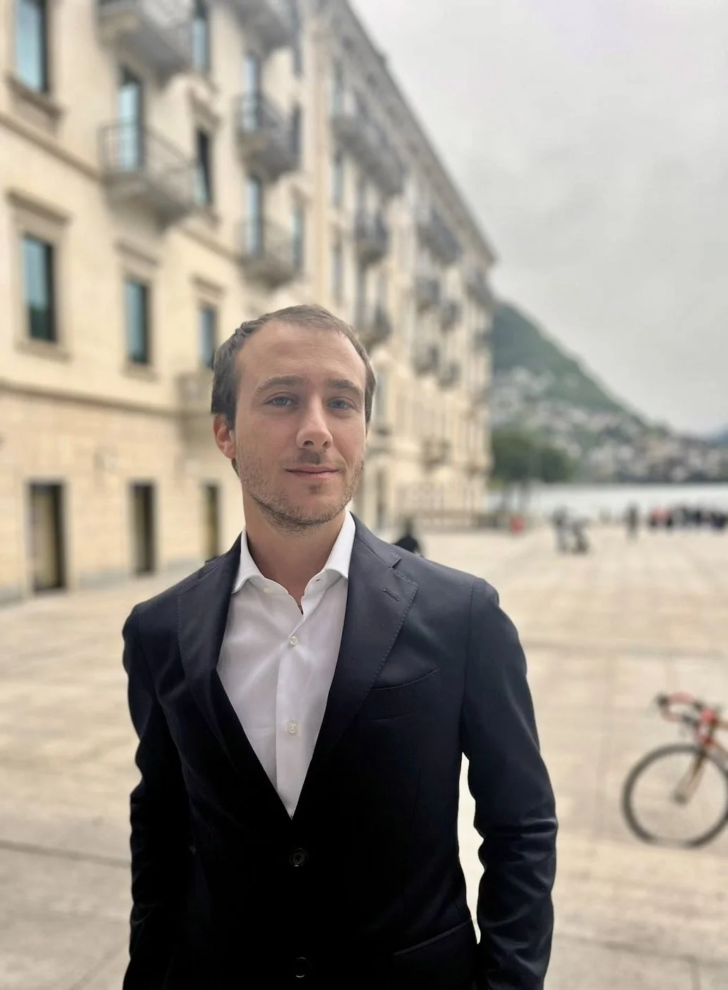 A well-dressed man in a black suit and white shirt standing outdoors by water with buildings and mountain in the background.