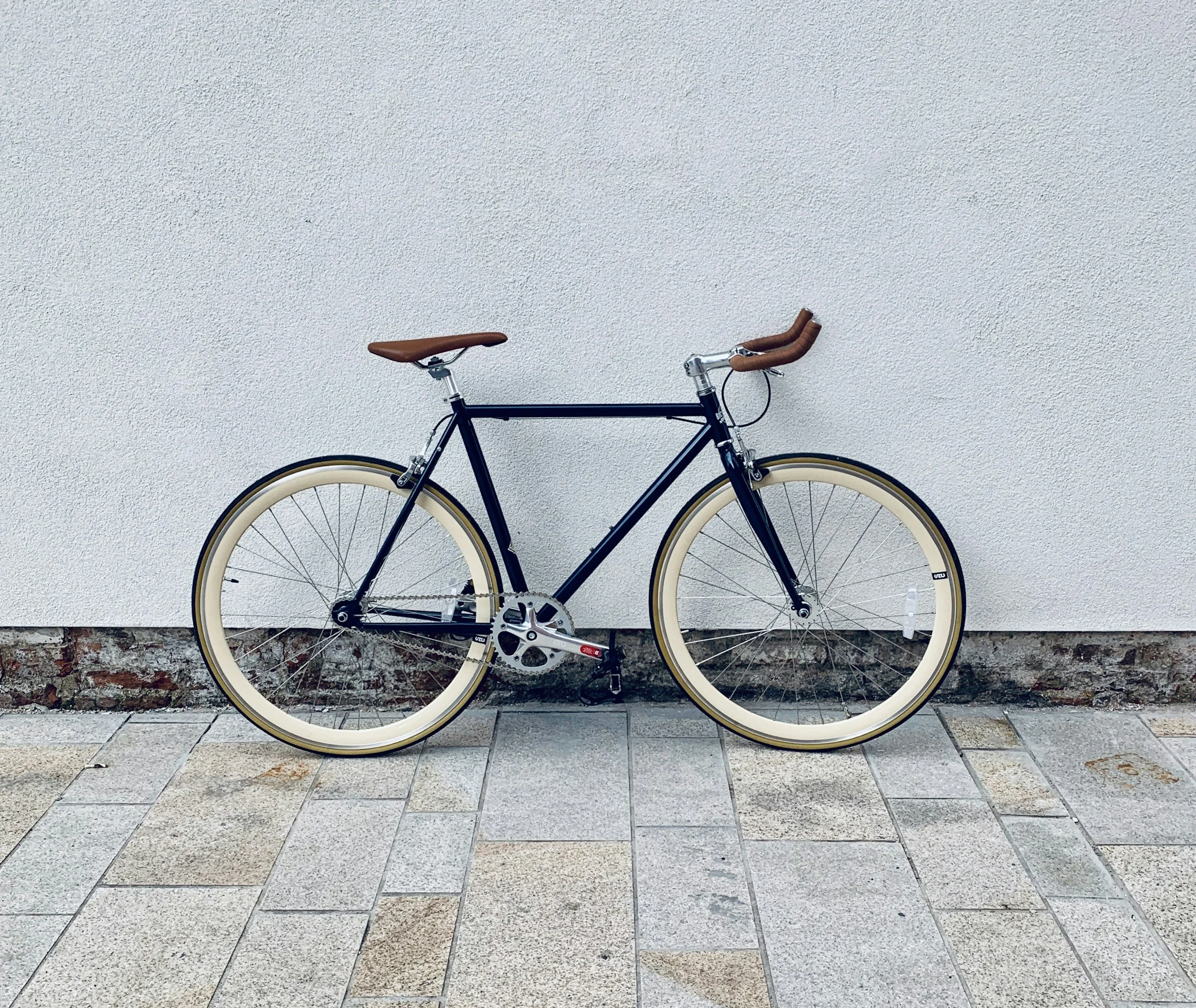 A black bicycle with brown handlebars and a brown seat, leaning against a white textured wall, on a paved sidewalk.