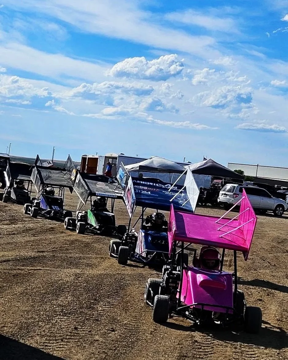 Miniature go-karts in a line on a dirt track, each with a colorful canopy, under a blue sky with clouds.