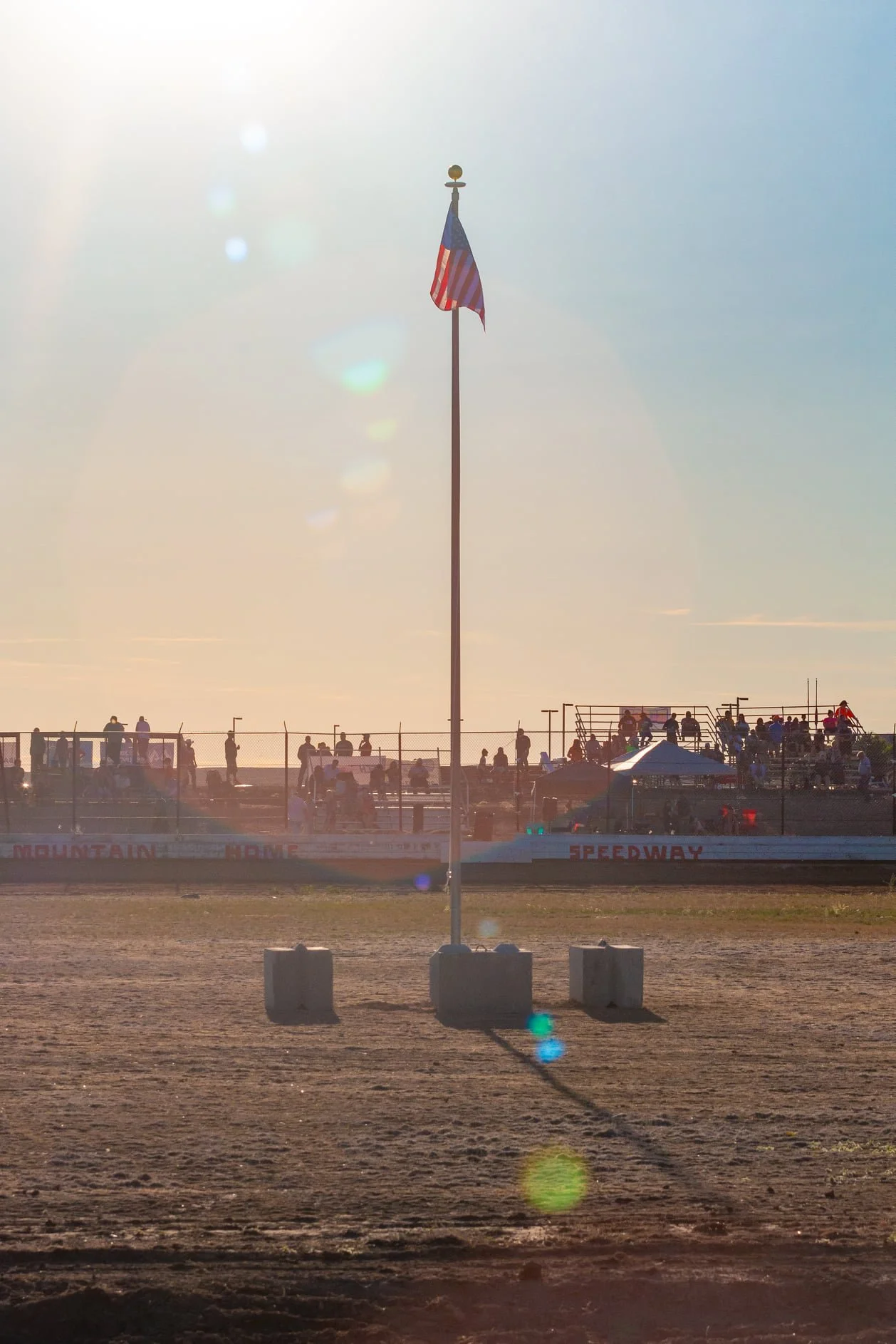 A flagpole with an American flag at a racing track during sunset, with spectators watching from the bleachers and the starting line in the foreground.